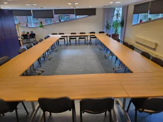 Een ruime vergaderruimte met een grote U-vormige tafelopstelling, omringd door zwarte stoelen. De kamer is licht en modern, met grote ramen die zorgen voor veel natuurlijk licht. Planten en decoratieve elementen voegen een frisse en uitnodigende sfeer toe. De ruimte is ideaal voor grote bijeenkomsten, presentaties of workshops.