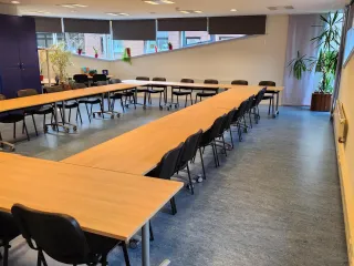 Een ruime vergaderruimte met een grote U-vormige tafelopstelling, omringd door zwarte stoelen. De kamer is licht en modern, met grote ramen die zorgen voor veel natuurlijk licht. Planten en decoratieve elementen voegen een frisse en uitnodigende sfeer toe. De ruimte is ideaal voor grote bijeenkomsten, presentaties of workshops.
