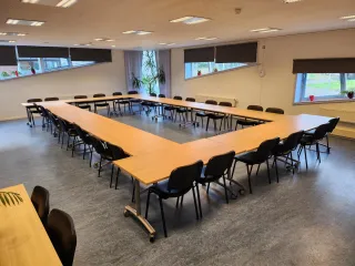 Een ruime vergaderruimte met een grote U-vormige tafelopstelling, omringd door zwarte stoelen. De kamer is licht en modern, met grote ramen die zorgen voor veel natuurlijk licht. Planten en decoratieve elementen voegen een frisse en uitnodigende sfeer toe. De ruimte is ideaal voor grote bijeenkomsten, presentaties of workshops.