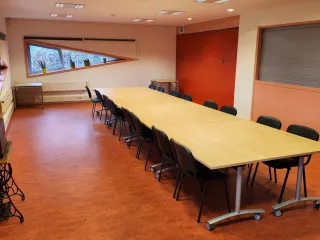 Een nette vergaderruimte met een lange houten tafel, omringd door zwarte stoelen. De schuine ramen met planten op de vensterbanken zorgen voor natuurlijk licht en een frisse uitstraling. De warme, rode vloer en opvallende rode kastwand geven de ruimte een levendige en functionele sfeer, ideaal voor formele bijeenkomsten of teamoverleg.