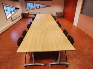 Een nette vergaderruimte met een lange houten tafel, omringd door zwarte stoelen. De schuine ramen met planten op de vensterbanken zorgen voor natuurlijk licht en een frisse uitstraling. De warme, rode vloer en opvallende rode kastwand geven de ruimte een levendige en functionele sfeer, ideaal voor formele bijeenkomsten of teamoverleg.