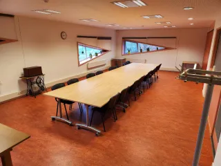 Een nette vergaderruimte met een lange houten tafel, omringd door zwarte stoelen. De schuine ramen met planten op de vensterbanken zorgen voor natuurlijk licht en een frisse uitstraling. De warme, rode vloer en opvallende rode kastwand geven de ruimte een levendige en functionele sfeer, ideaal voor formele bijeenkomsten of teamoverleg.