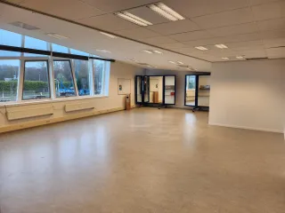 Een ruime, open zaal met grote ramen die veel natuurlijk licht binnenlaten en uitzicht bieden op een buitenomgeving. De lichte vloer en het strakke, witte plafond creëren een frisse en veelzijdige ruimte, ideaal voor activiteiten zoals vergaderingen, workshops of bewegingslessen. Verplaatsbare spiegels aan de achterkant voegen flexibiliteit toe aan de inrichting, terwijl de moderne uitstraling de ruimte functioneel en uitnodigend maakt.