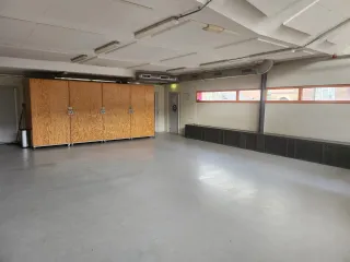 Een ruime multifunctionele zaal met een strak en eenvoudig ontwerp. Grote houten opbergkasten bieden praktische opslagmogelijkheden, terwijl de lange ramen voor natuurlijk licht zorgen. De open indeling maakt de ruimte flexibel en geschikt voor verschillende activiteiten zoals workshops, bijeenkomsten of sportlessen.