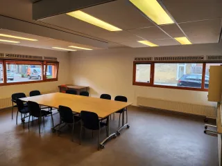 Een ruime vergaderruimte met een grote houten tafel, omringd door blauwe stoelen. Grote ramen zorgen voor natuurlijk licht en een open sfeer, terwijl ingebouwde verlichting de ruimte helder en functioneel maakt.