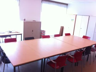 Een lichte en functionele vergaderruimte met een grote, rechthoekige tafel omringd door rode en zwarte stoelen. Grote ramen met witte gordijnen zorgen voor veel natuurlijk licht en een open sfeer. Aan de zijkant staat een opbergkast en extra tafels, wat de ruimte praktisch en veelzijdig maakt. De minimalistische inrichting maakt het ideaal voor vergaderingen, workshops of kleine bijeenkomsten.