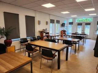 De huiskamer in het Buitenhuis is een lichte en gezellige ruimte met houten tafels, comfortabele stoelen en grote ramen. Voorzien van een televisie, boekenplank en directe toegang tot buiten, ideaal voor diverse activiteiten en bijeenkomsten.