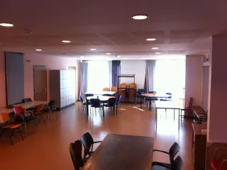 Een veelzijdige en ruime zaal met verschillende tafels en stoelen, ideaal voor groepsactiviteiten of bijeenkomsten. Grote ramen met gordijnen laten veel natuurlijk licht binnen, wat de ruimte open en uitnodigend maakt. Een rij kluisjes staat langs de muur voor praktische opslag, terwijl extra stoelen en een kapstok achterin de zaal flexibiliteit bieden. De warme tinten van de vloer en het meubilair creëren een comfortabele en functionele sfeer.