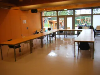 Een ruime zaal met lange tafels in een flexibele opstelling, ideaal voor vergaderingen of workshops. Grote ramen en glazen deuren bieden veel lichtinval en zicht op een buitenspeelplaats. De warme muurkleuren en houten accenten creëren een aangename, uitnodigende sfeer.