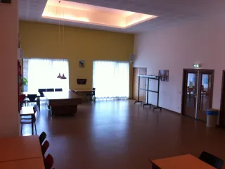 Een ruime en sfeervolle zaal met een hoge gele accentmuur en grote ramen voorzien van lichte gordijnen, die veel natuurlijk licht binnenlaten. In het midden staat een biljarttafel, wat de ruimte een sociale en multifunctionele functie geeft. Hangende lampen voegen een warm accent toe, terwijl tafels, stoelen en een verrijdbare kapstok aan de zijkant de zaal flexibel en praktisch in gebruik maken. Ideaal voor bijeenkomsten, ontspanning of evenementen.