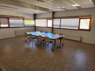 Een lichte en open activiteitenruimte met grote ramen die natuurlijk licht binnenlaten. De ruimte is voorzien van een centrale tafel met stoelen, geschikt voor vergaderingen, workshops of kleine groepsactiviteiten. Het eenvoudige interieur en de flexibele indeling maken het een veelzijdige plek voor diverse doeleinden.