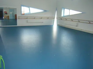 Een lichte en ruime oefenruimte met een blauwe vloer en strakke witte muren. De grote spiegels aan de ene kant en de houten barre langs de muren maken de zaal perfect voor dans- of bewegingslessen. De driehoekige ramen zorgen voor natuurlijk licht en geven de ruimte een moderne uitstraling.