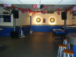  Deze jongerenruimte heeft een moderne, feestelijke uitstraling met blauwe muren en rode en roze versieringen aan het plafond. In het midden hangen twee verlichte dartborden met een scorebord ertussen, perfect voor een potje darten. Links staat een Red Bull-koeler, terwijl rechts een voetbaltafel en hoge krukken rond een bar zichtbaar zijn. Grote speakers en spotlampen wijzen op mogelijkheden voor muziek en feestjes. De ruimte straalt gezelligheid uit en biedt plek voor ontspanning, spel en samenzijn.
