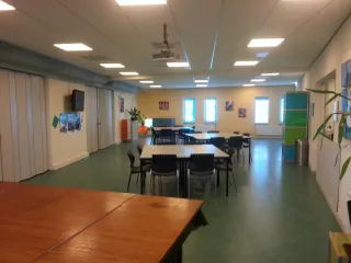 De grote zaal in het Buitenhuis is een veelzijdige ruimte die ideaal is voor grotere bijeenkomsten, evenementen of groepsactiviteiten. De zaal heeft een flexibele opstelling met tafels en stoelen die gemakkelijk aanpasbaar zijn aan verschillende behoeften. Het interieur is licht en ruim, met voldoende ramen die natuurlijk licht binnenlaten. Daarnaast zijn er moderne voorzieningen, zoals een projector en een televisie, waardoor deze ruimte uitstekend geschikt is voor presentaties en workshops.