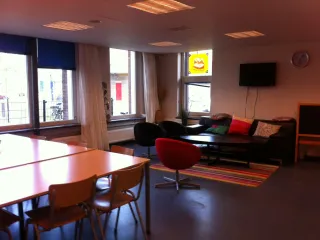 Een gezellige huiskamerruimte met een uitnodigende zithoek bestaande uit een bank en kleurrijke kussens, een laag salontafeltje en comfortabele fauteuils. De ruimte heeft een warme sfeer dankzij de houten tafels, stoelen en het kleurrijke tapijt. Grote ramen met gordijnen zorgen voor veel natuurlijk licht, terwijl een muur met een tv en een krijtbord de functionaliteit versterken. Deze ruimte is ideaal voor informele bijeenkomsten, ontspanning of sociale activiteiten.