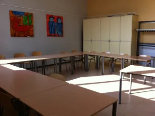 Een lichte en ruime activiteitenruimte met een U-vormige tafelopstelling en houten stoelen eromheen, ideaal voor vergaderingen of groepsactiviteiten. De hoge kasten aan de achterkant bieden praktische opbergruimte, terwijl kleurrijke schilderijen aan de muur de ruimte een creatieve en levendige uitstraling geven. De grote ramen zorgen voor veel natuurlijk licht, wat de ruimte uitnodigend en prettig maakt.