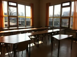 Een lichte les- of activiteitenruimte met grote ramen die een panoramisch uitzicht bieden en zorgen voor een overvloed aan natuurlijk licht. De tafels en stoelen zijn georganiseerd in een flexibele opstelling, ideaal voor onderwijs of groepswerk. De zachte oranje gordijnen voegen een warme en uitnodigende sfeer toe. De ruimte heeft een praktische, functionele uitstraling, geschikt voor diverse activiteiten.
