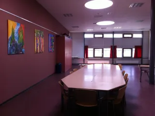 Een stijlvolle zaal met een warme, donkerrode accentmuur, voorzien van kleurrijke kunstwerken die de ruimte een creatieve uitstraling geven. Grote ramen met rode gordijnen zorgen voor natuurlijk licht en bieden een warme sfeer. Een lange tafel met houten stoelen in het midden maakt de ruimte ideaal voor vergaderingen of groepsactiviteiten. De plafondverlichting, draagt bij aan een moderne en goed verlichte omgeving, terwijl de hoge kast aan de zijkant zorgt voor praktische opbergruimte.