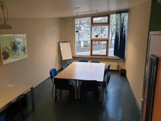 Een compacte en functionele vergaderruimte met een eenvoudige opstelling van tafels en blauwe stoelen, geschikt voor kleine groepen. Het grote raam laat veel natuurlijk licht binnen en biedt uitzicht op een rustige straat. Aan de muur hangt een kunstwerk dat kleur en persoonlijkheid toevoegt aan de ruimte. Een flip-over in de hoek benadrukt de praktische bruikbaarheid van de ruimte voor brainstormsessies of presentaties. De kamer heeft een rustige, professionele uitstraling.