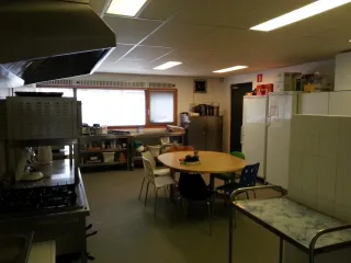 Een volledig uitgeruste keuken met een praktische indeling. Voorzien van een groot fornuis, opslagrekken en meerdere koelkasten. In het midden staat een ronde tafel met stoelen, ideaal voor informele bijeenkomsten of voorbereiding. De ruimte is functioneel en goed verlicht, geschikt voor kookactiviteiten of catering.
